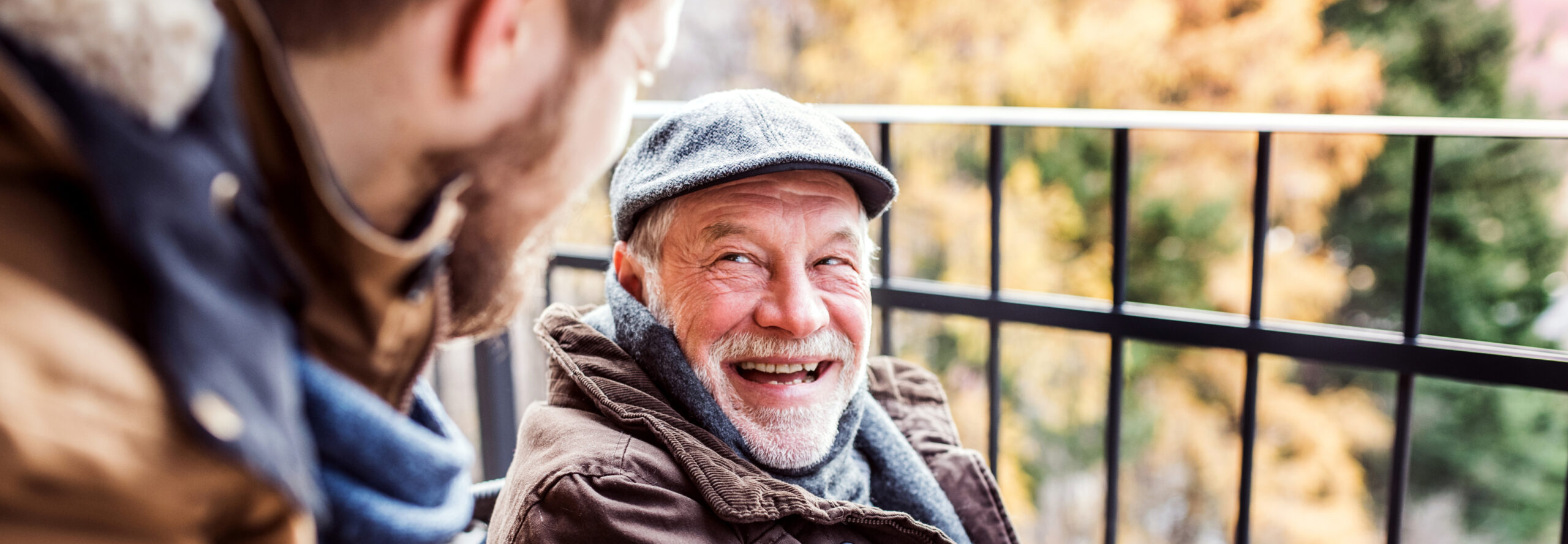 Older gentleman wearing a hat and coat outside laughs with yonger gentleman